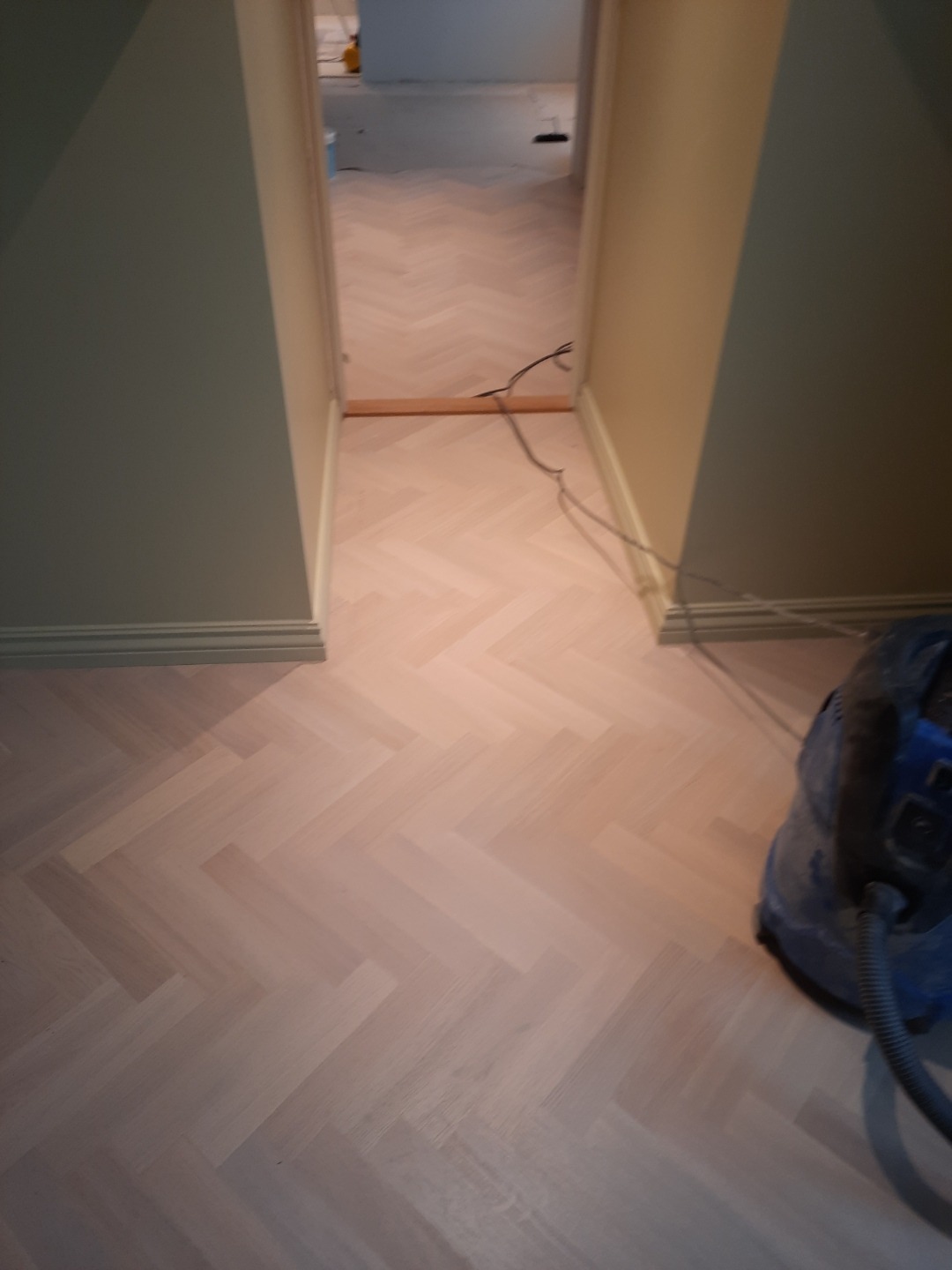 Jasna, parkietowa podłoga ułożona w jodełkę w korytarzu, z widocznym odkurzaczem przemysłowym w rogu. Ściany w odcieniach beżu i szarości. Widok w głąb pomieszczenia.