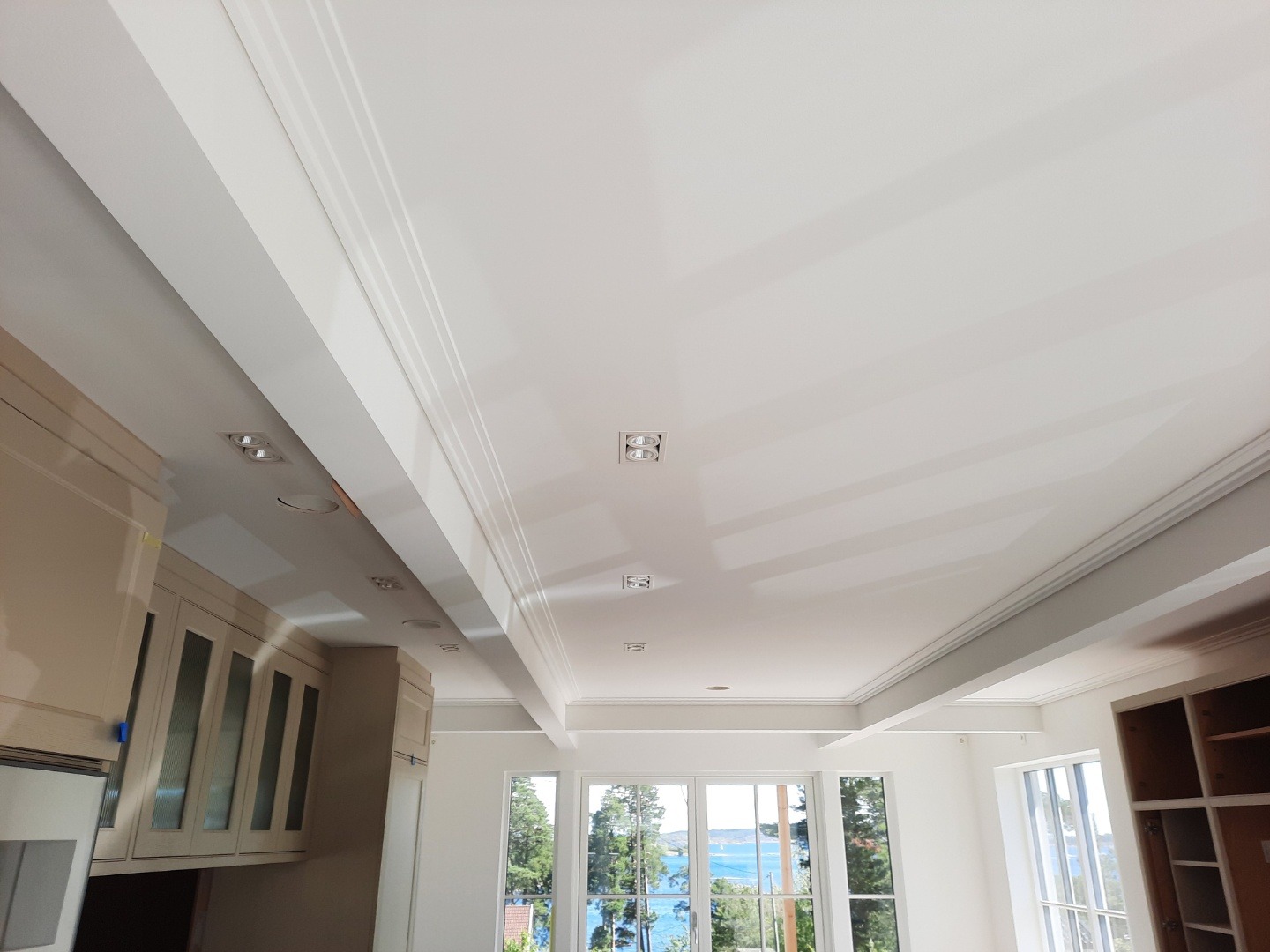 Jasne wnętrze z widokiem na morze przez okna. Sufit z dekoracyjnymi listwami i wbudowanym oświetleniem. Częściowo widoczne szafki kuchenne w neutralnych barwach.