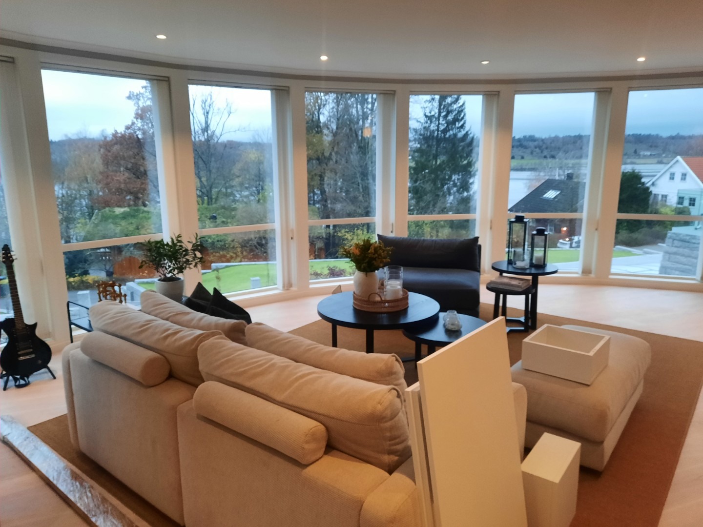 Jasny salon z panoramicznymi oknami, beżową sofą i czarną gitarą elektryczną w rogu. Widok na zieleń za oknem. Minimalistyczny wystrój.