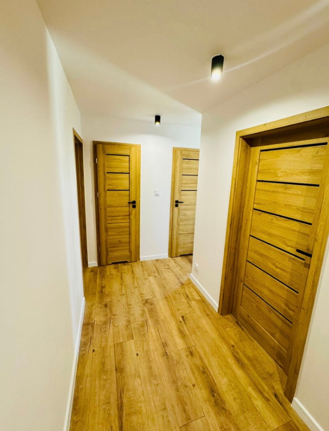 Korytarz z trzema drewnianymi drzwiami z poziomymi panelami i czarnymi klamkami, oświetlony dwoma punktowymi lampami sufitowymi, z podłogą imitującą drewno.
