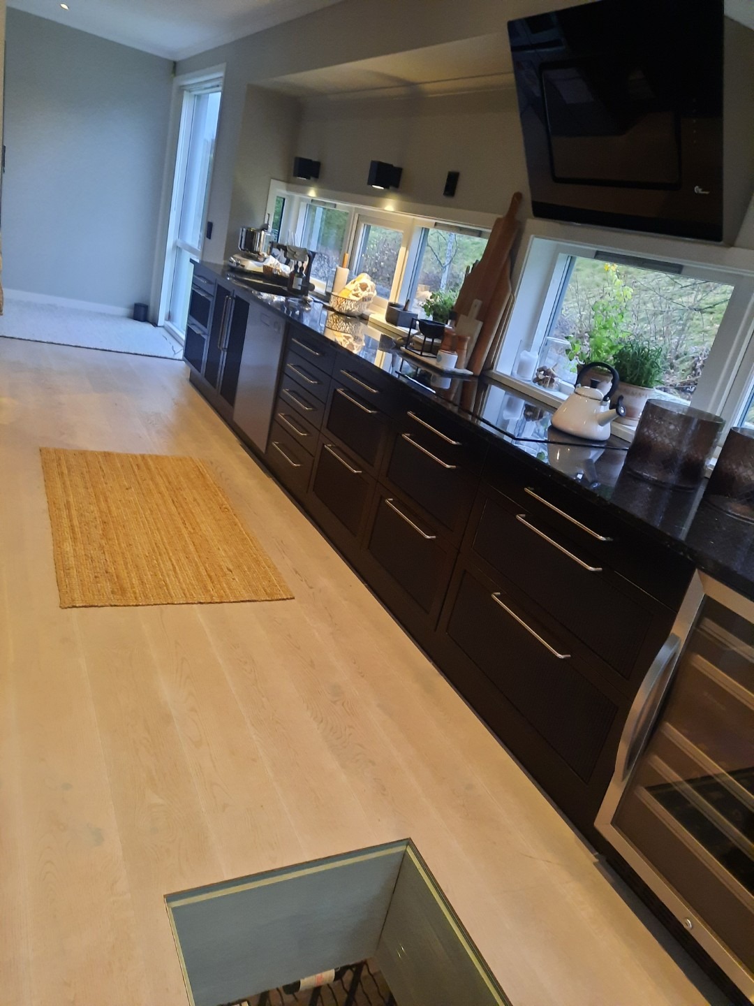 Nowoczesna kuchnia z czarnymi szafkami, blatem z ciemnego kamienia i jasną drewnianą podłogą. Widoczne okno z widokiem na zieleń i dywanik na podłodze.