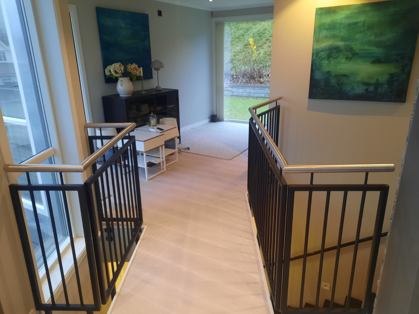 Nowoczesne wnętrze z czarną balustradą schodową i jasną podłogą. W tle widoczne obrazy i okno z widokiem na zieleń. Minimalistyczny design.