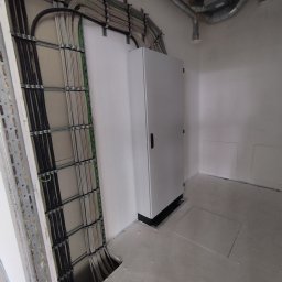 Pabian instalacje elektryczne - Szara szafa elektryczna na tle ściany z widocznymi kablami w metalowych korytach. Dodatkowo widoczne rury wentylacyjne pod sufitem. Surowy stan.