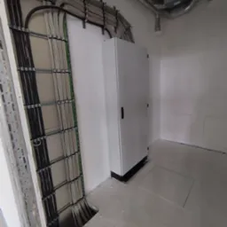 Szara szafa elektryczna na tle ściany z widocznymi kablami w metalowych korytach. Dodatkowo widoczne rury wentylacyjne pod sufitem. Surowy stan.