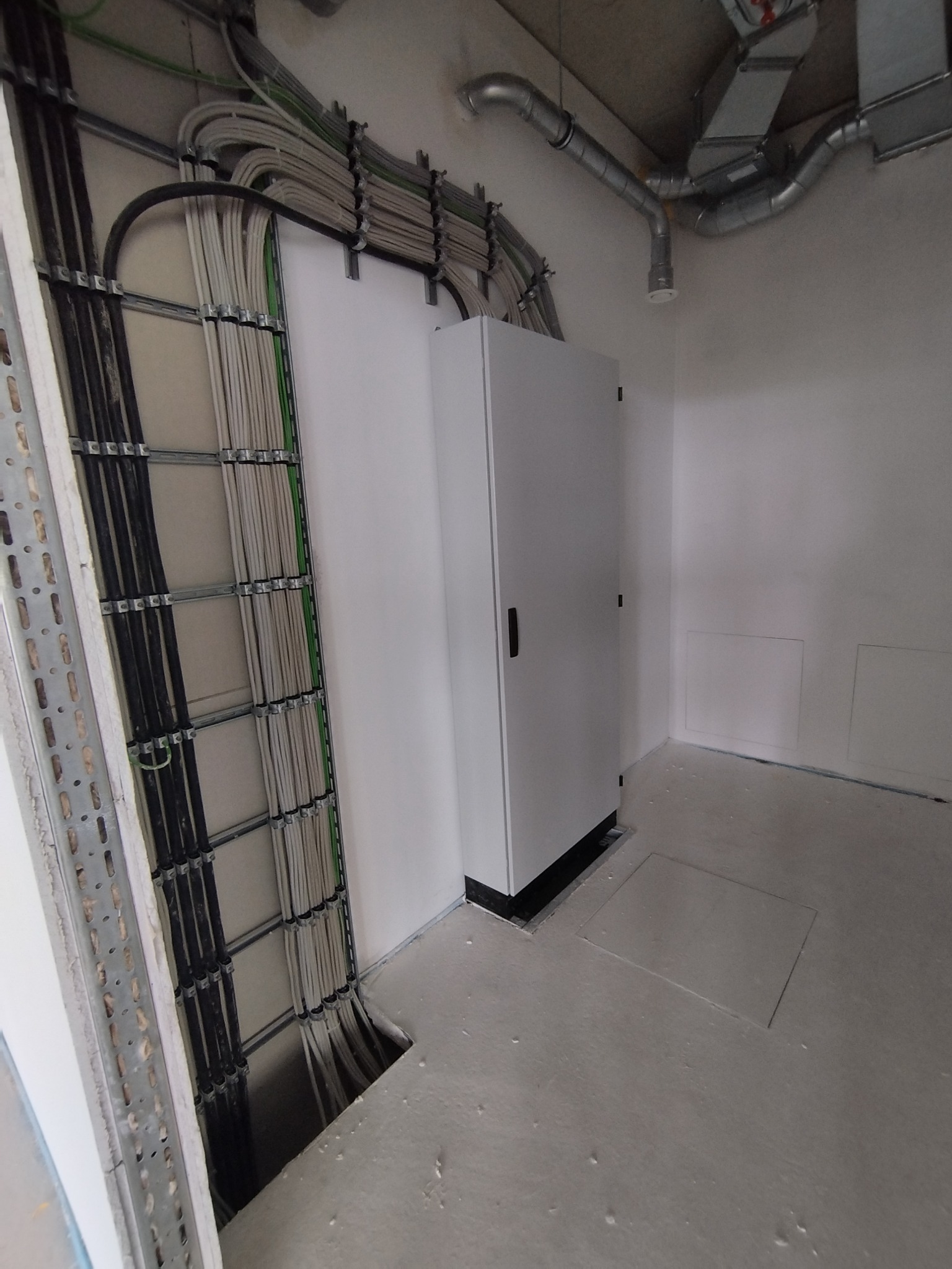 Szara szafa elektryczna na tle ściany z widocznymi kablami w metalowych korytach. Dodatkowo widoczne rury wentylacyjne pod sufitem. Surowy stan.