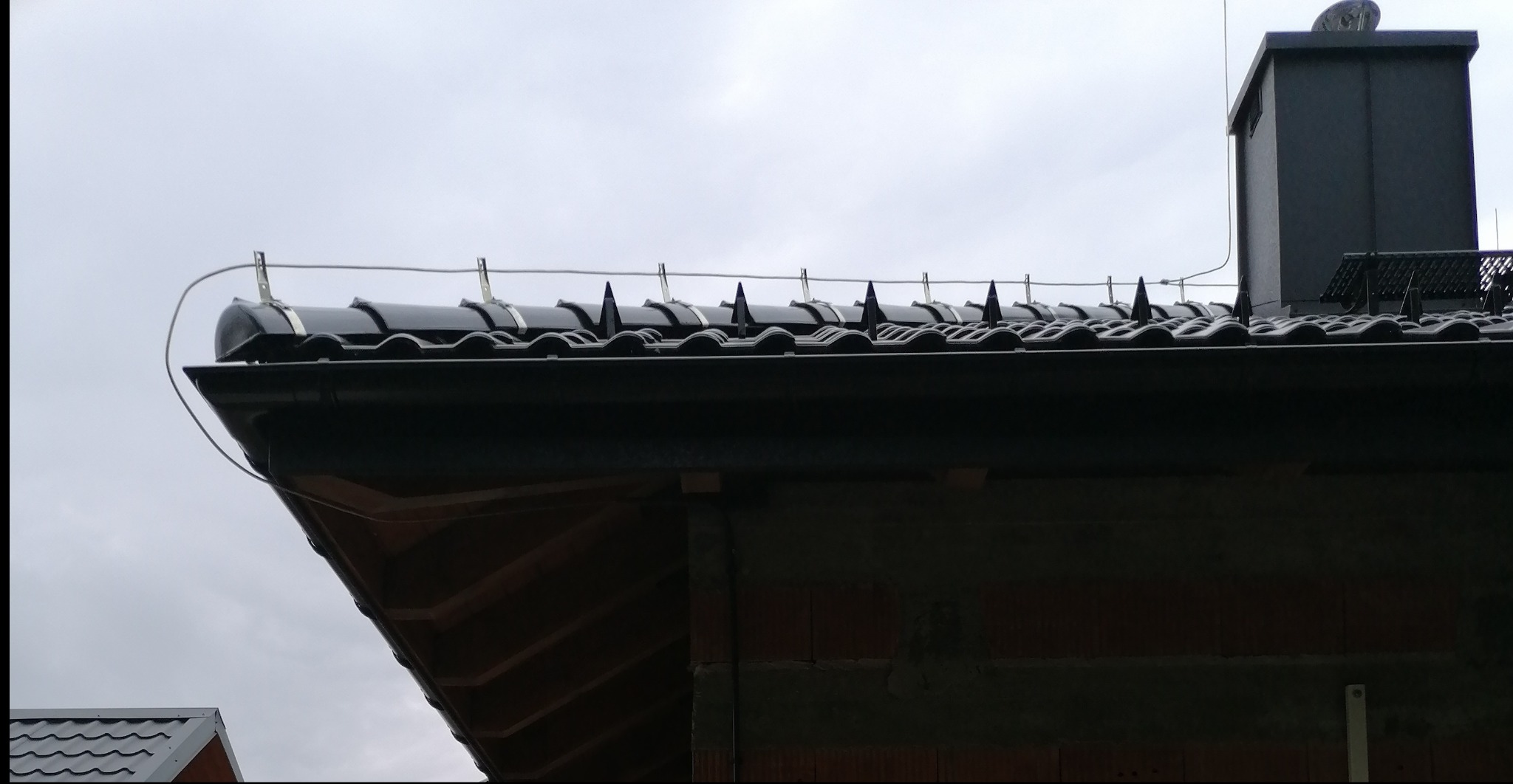 Czarny dach z zamontowaną instalacją odgromową z metalowymi uchwytami i przewodem, widok z dołu na tle pochmurnego nieba.