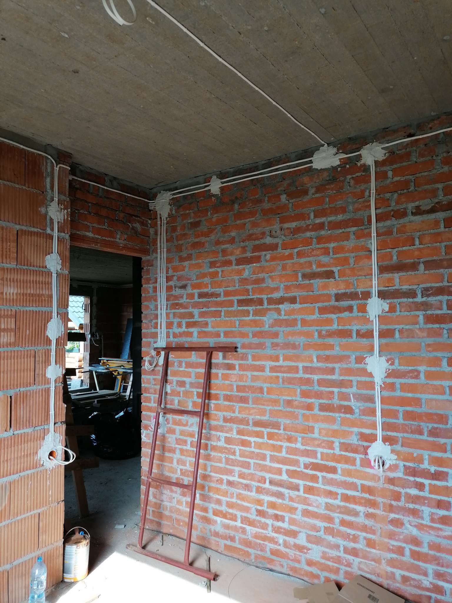 Surowy mur z instalacją elektryczną w peszlach, zabezpieczoną gipsem. Widoczna drabina malarska i fragment pomieszczenia w stanie deweloperskim.