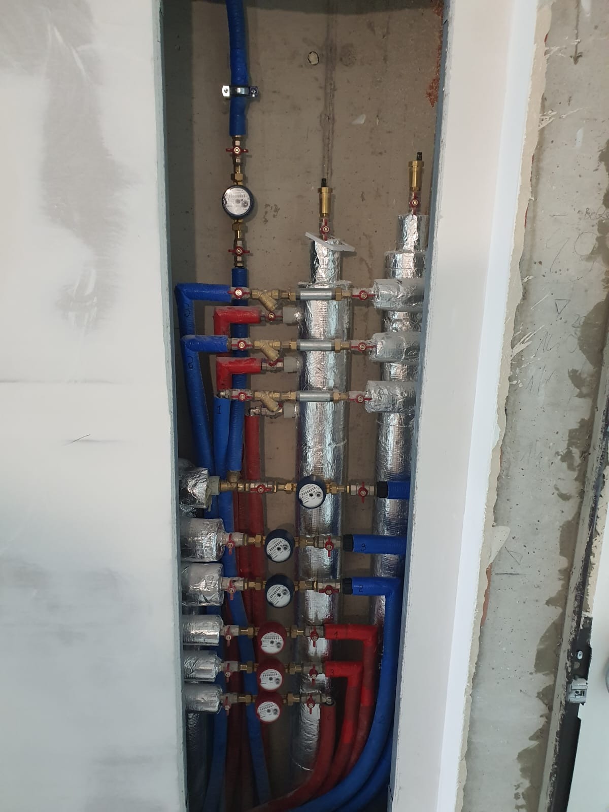 Rozdzielacz ogrzewania podłogowego z izolowanymi rurami, zaworami i wodomierzami, zamontowany w surowym otworze ściennym. Instalacja grzewcza w trakcie prac wykończeniowych.