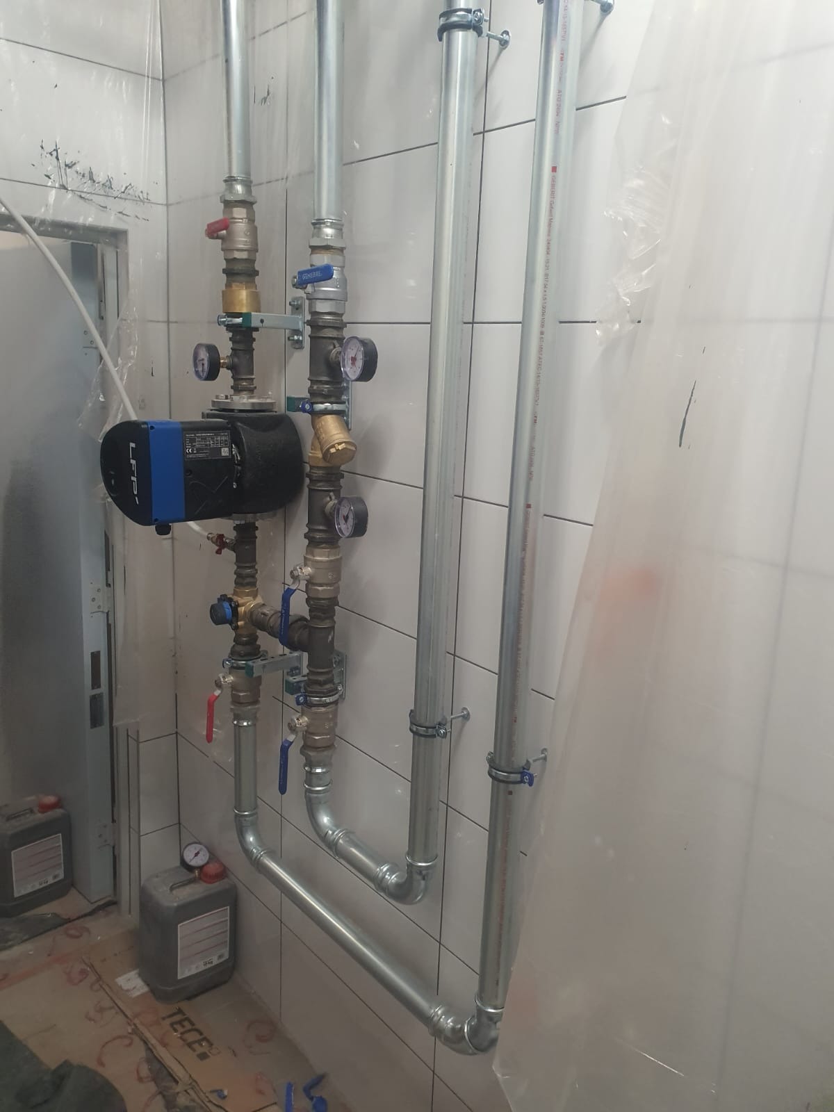 Nowa instalacja hydrauliczna z pompą obiegową LFP, rurami stalowymi i zaworami, zamontowana na ścianie z białych płytek w pomieszczeniu w trakcie prac wykończeniowych.
