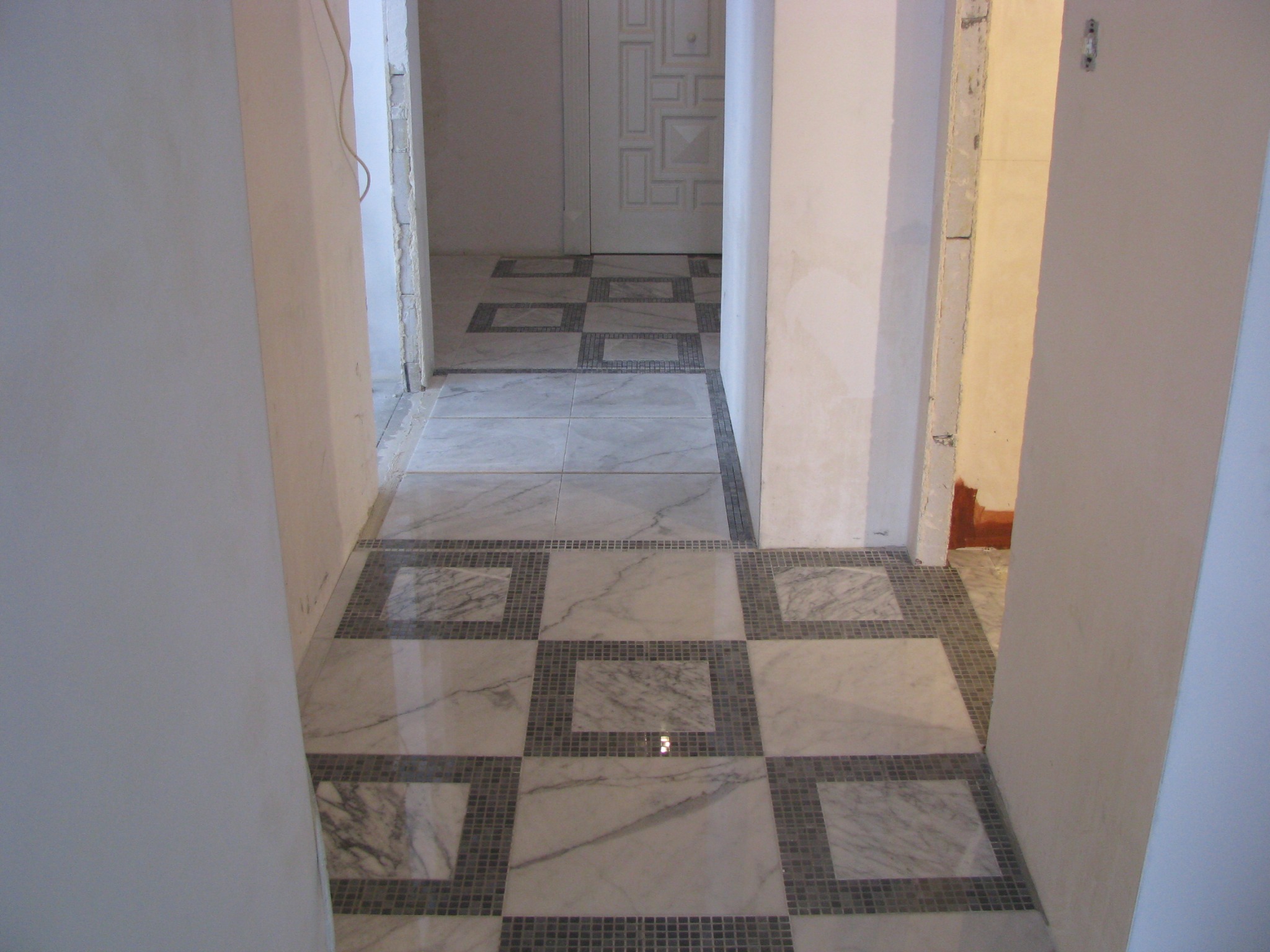 Korytarz z marmurową posadzką w biało-szarej tonacji, ułożoną we wzór szachownicy z mozaikowymi obramowaniami. Widoczne surowe ściany i futryny.
