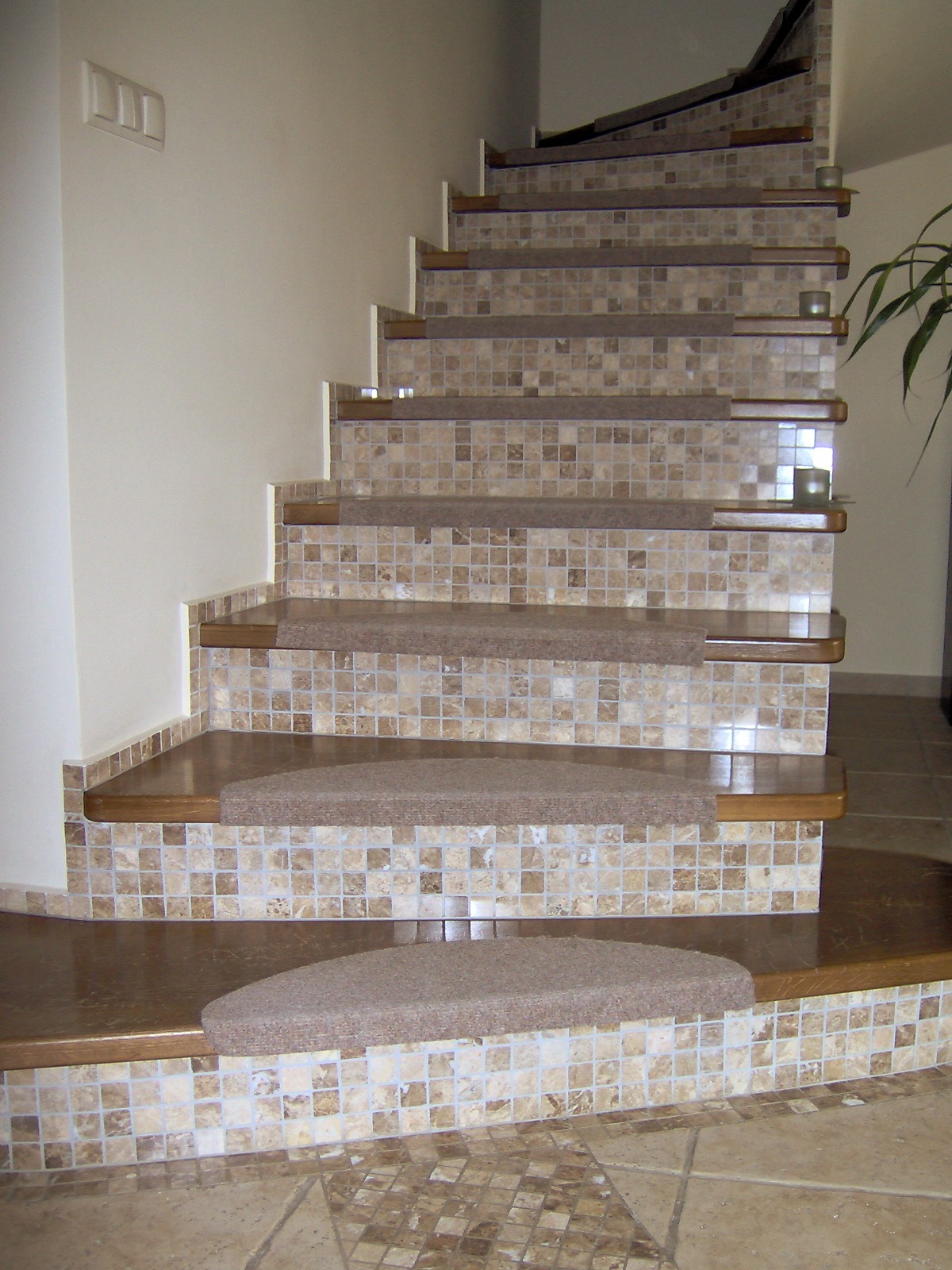 Wykończone schody z mozaiką w odcieniach beżu i brązu, z drewnianymi krawędziami i nakładkami dywanowymi. Widoczne detale wykończenia i wzór mozaiki na podłodze.