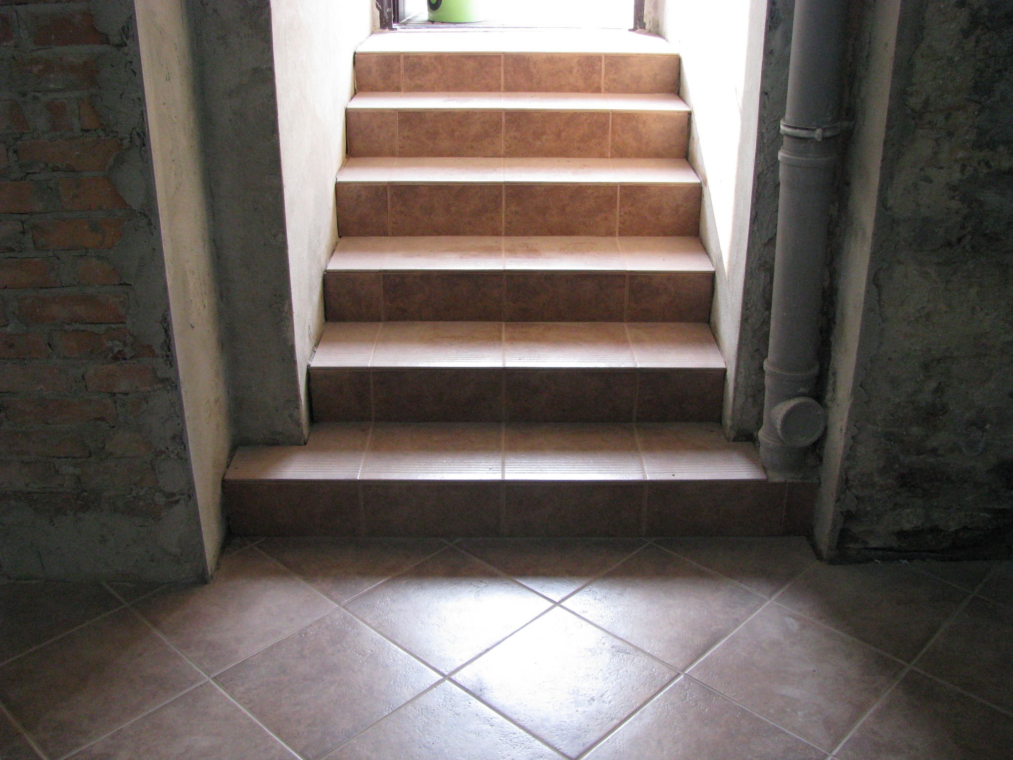 Schody wyłożone płytkami w odcieniach beżu, prowadzące w górę. Na ścianie widoczna rura. Podłoga również wyłożona płytkami w romby. Surowe wykończenie ścian.