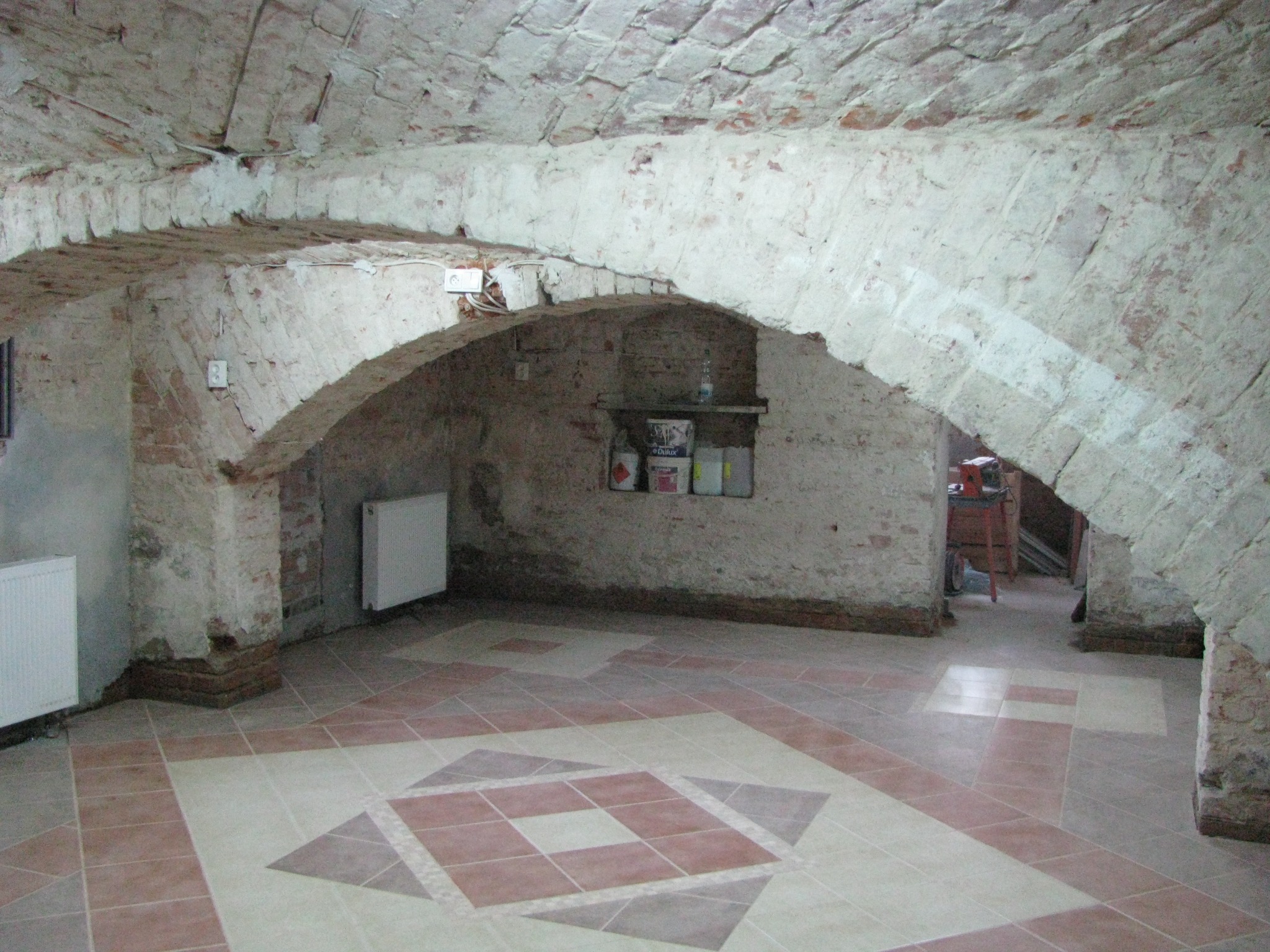 Klimatyczne wnętrze piwnicy z łukowym sklepieniem, wykończone płytkami podłogowymi w geometryczny wzór. Widoczne grzejniki i wnęka z półką.