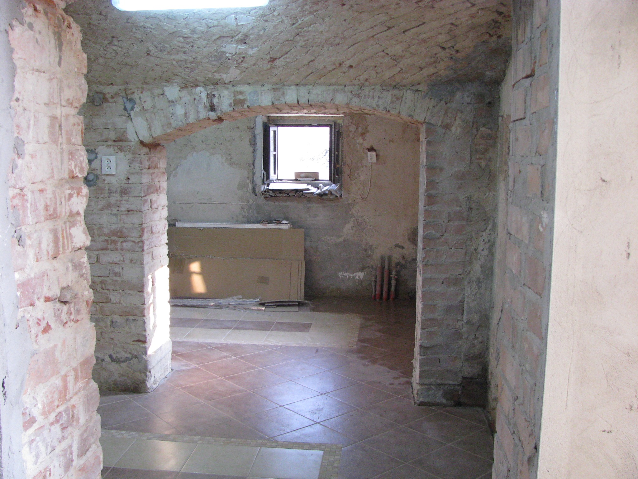 Wnętrze w trakcie remontu: surowe ceglane ściany z łukowym przejściem, częściowo ułożona terakota i okno z widokiem na zewnątrz, widoczne instalacje.