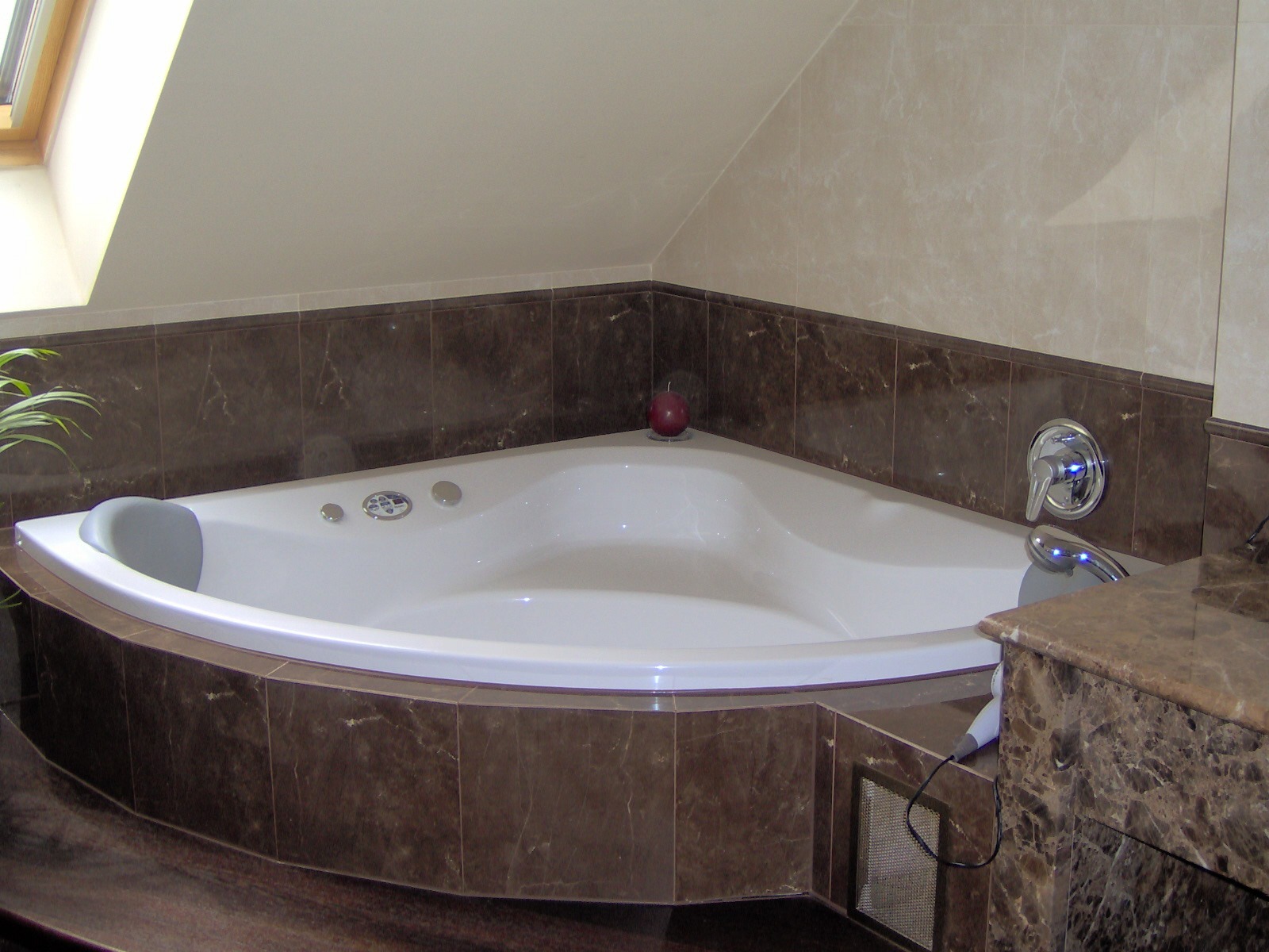 Biała, narożna wanna z hydromasażem, obudowana brązowymi płytkami imitującymi marmur. Na wannie dekoracyjna, czerwona kula. U góry okno dachowe.