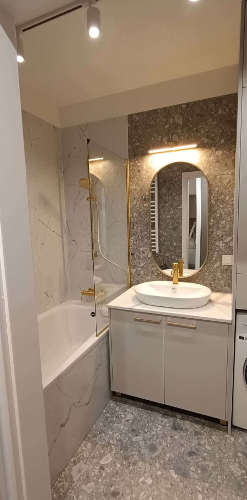 Nowoczesna łazienka z wanną, szklanym parawanem, umywalką na szafce i lustrem w złotej ramie. Na ścianach i podłodze płytki imitujące kamień.