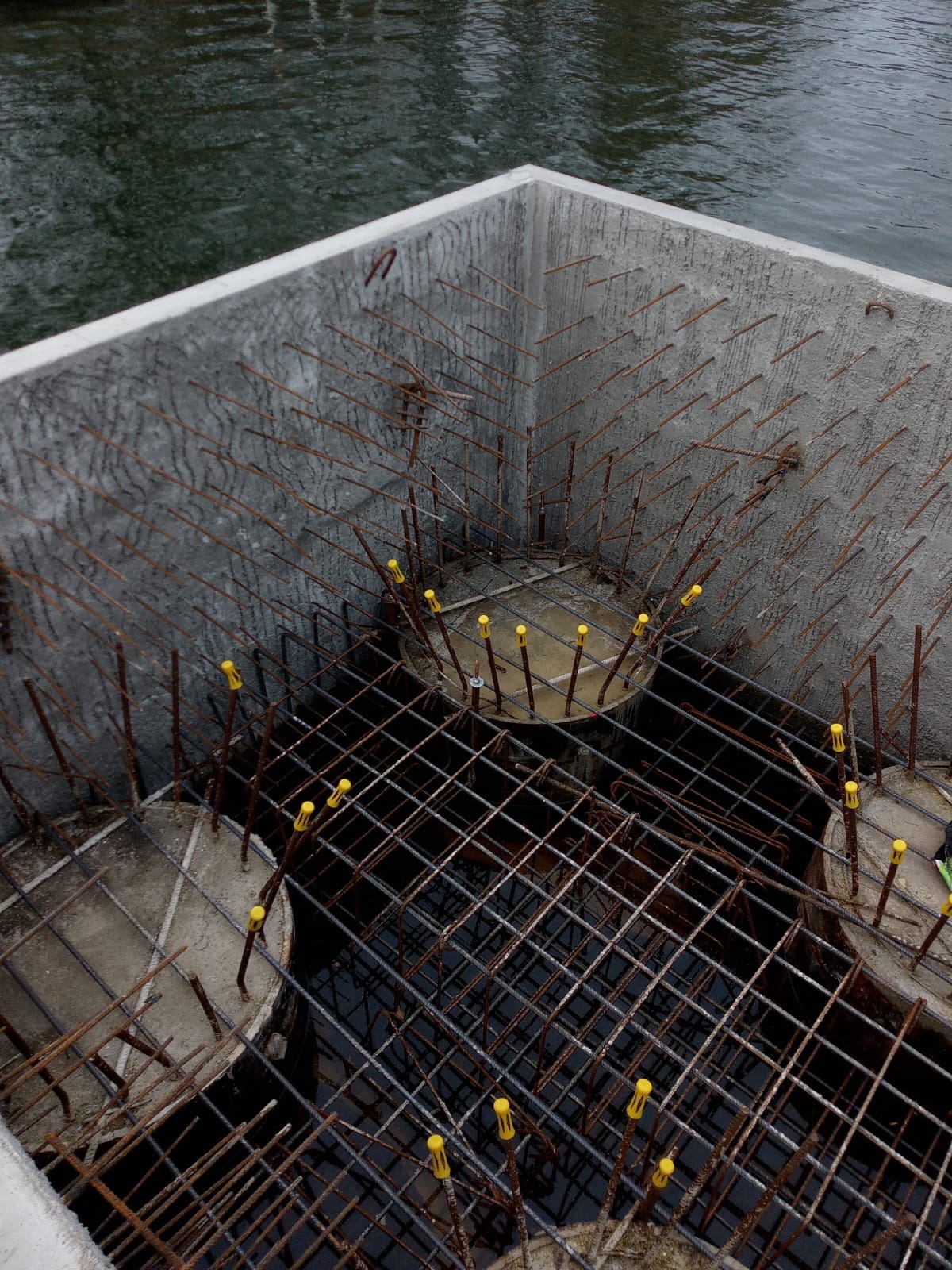 Zbrojenie betonowej konstrukcji z wystającymi prętami i żółtymi zabezpieczeniami, widok z góry na mokrą konstrukcję. W tle woda.