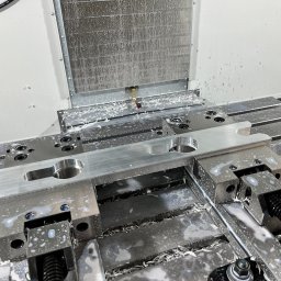 MIT-TECH - Metalowy detal w trakcie obróbki na frezarce CNC, widoczne wióry i chłodziwo. Precyzyjna praca na maszynie, detale mocowane w uchwytach.