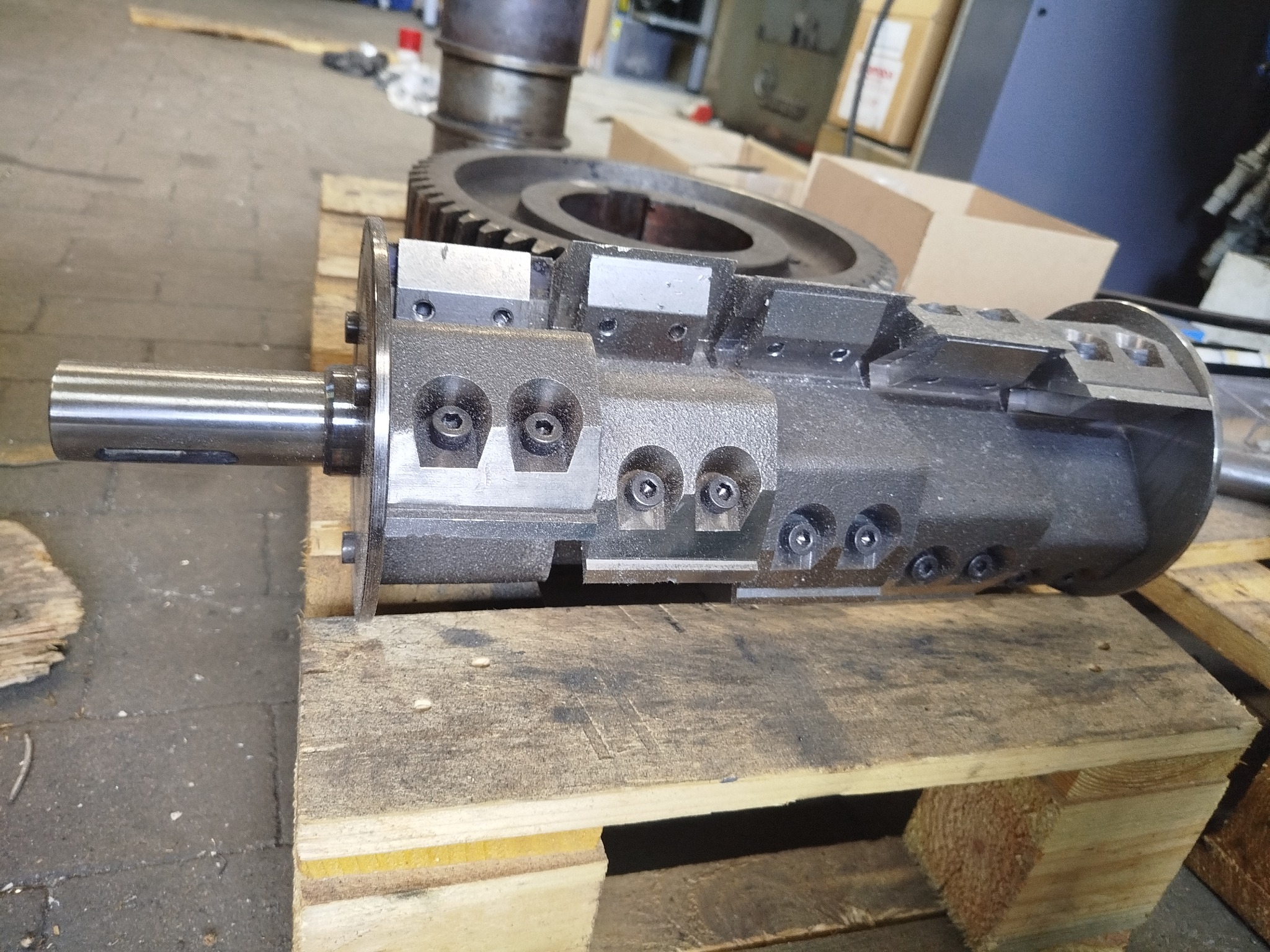 Szary, metalowy wał frezarski z ostrzami, leżący na drewnianej palecie. W tle widoczne elementy maszyny i przekładnia zębata.