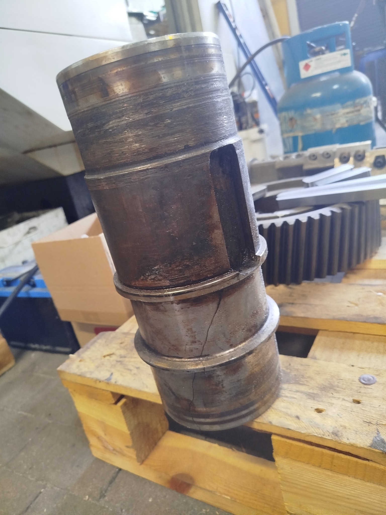 Uszkodzony, metalowy cylinder z pęknięciem, stojący na drewnianej palecie, zębatka i butla gazowa w tle. Widoczne ślady obróbki.