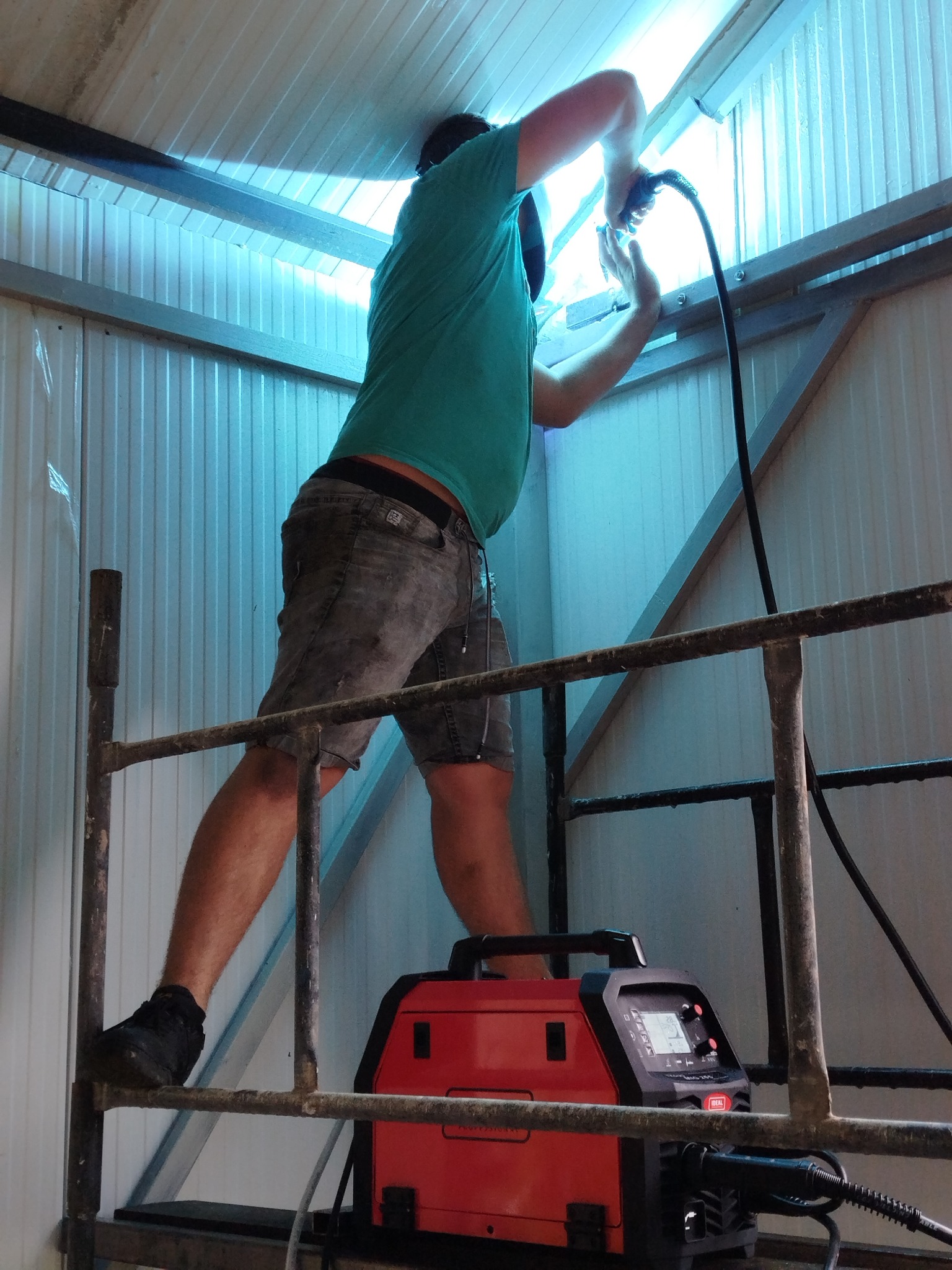 Spawacz na rusztowaniu łączy metalową konstrukcję wewnątrz budynku. Widoczny spawacz w szortach, spawarka z przewodem, metalowe elementy konstrukcji.