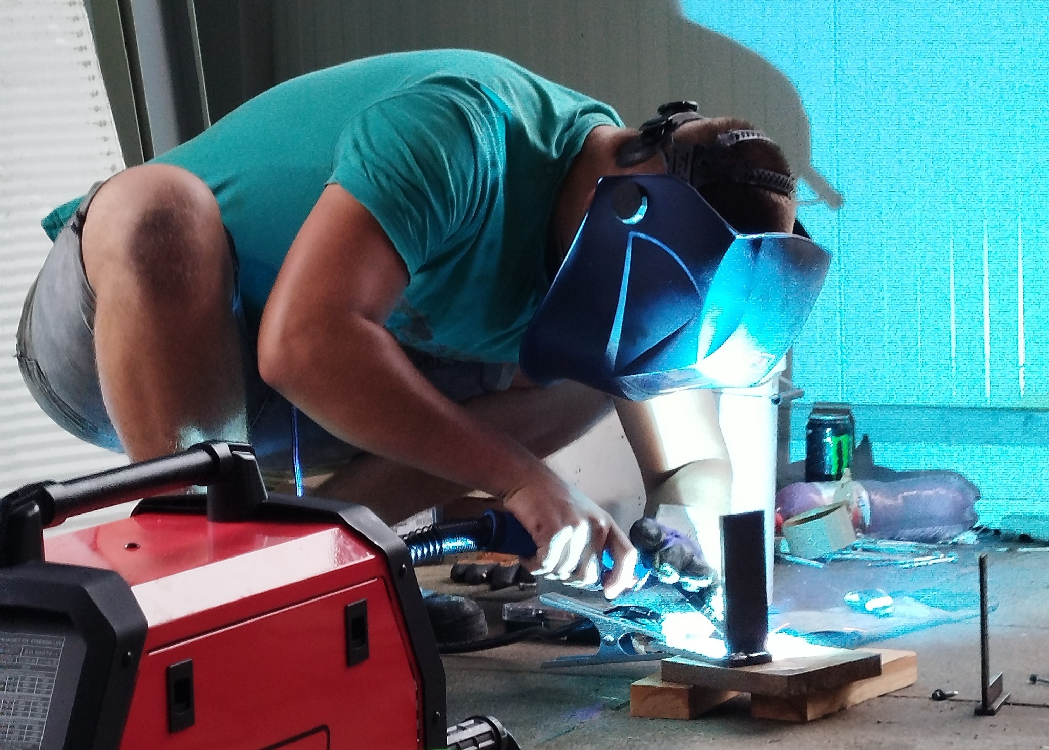 Pracownik w masce spawalniczej, w pozycji kucznej, spawa metalowe elementy na warsztacie. Czerwona spawarka widoczna w tle.