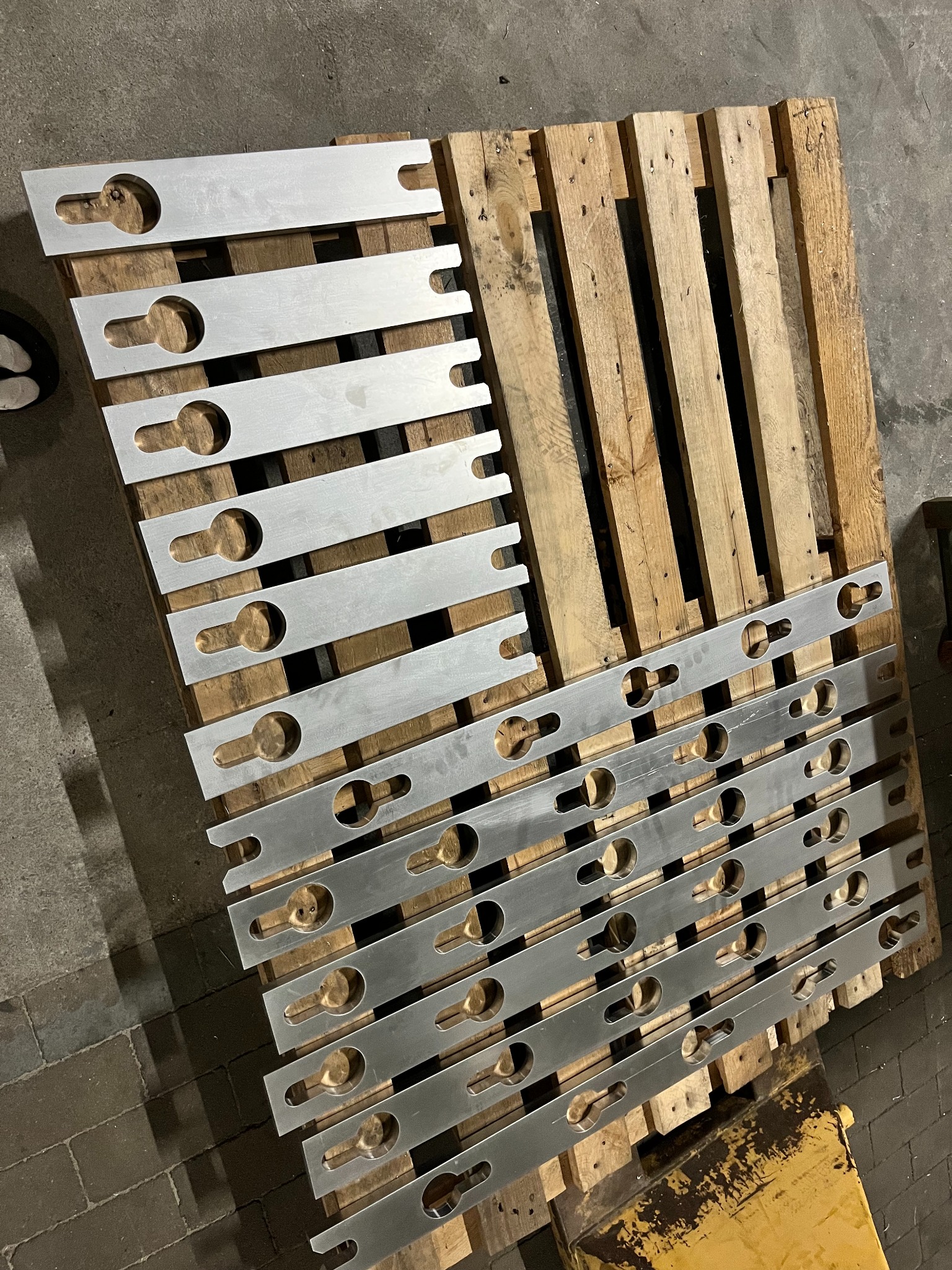 Metalowe elementy z otworami i wycięciami, ułożone na drewnianej palecie. Detal obróbki metalu, widoczne ślady po procesie produkcyjnym.