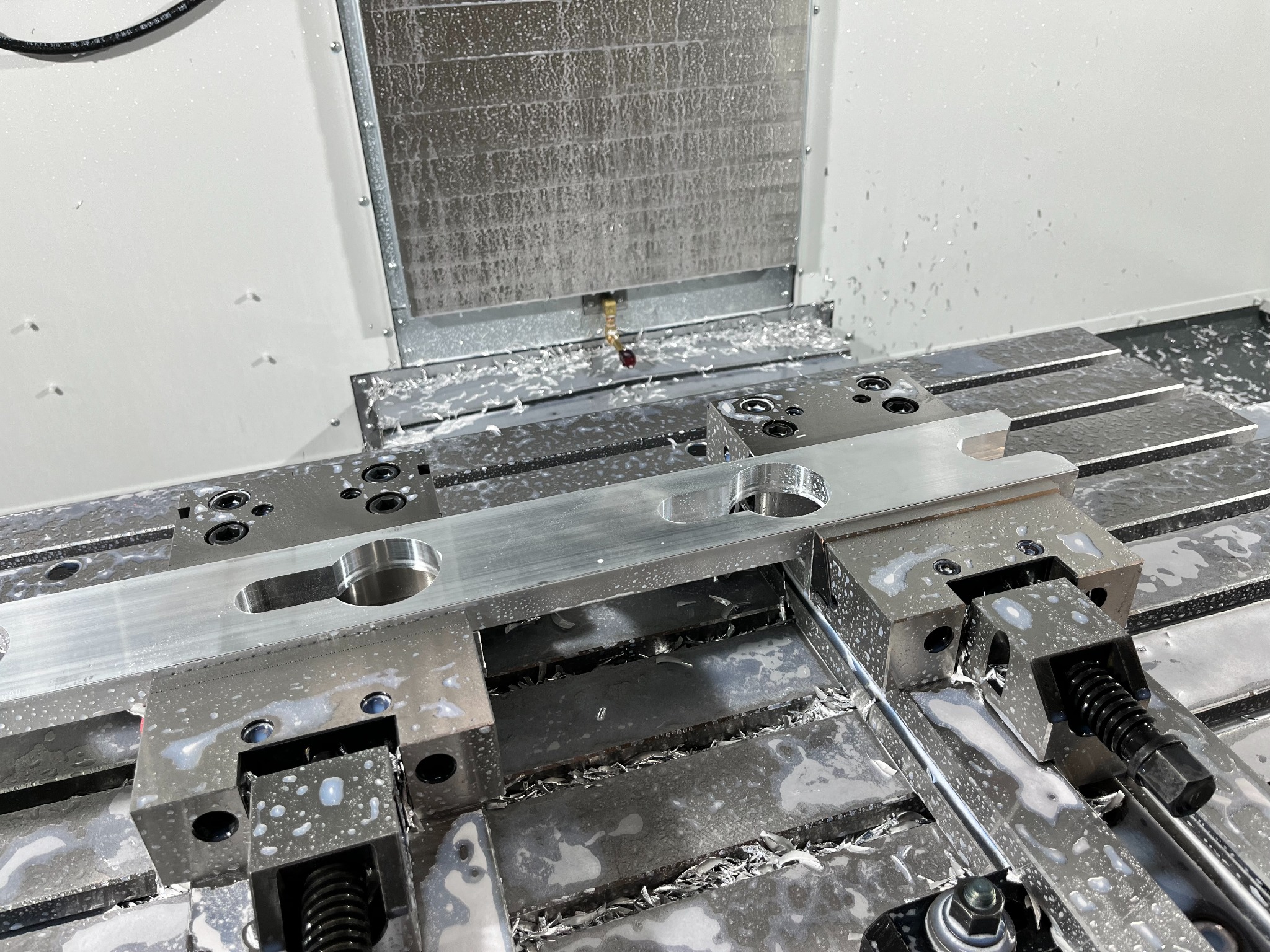 Metalowy detal w trakcie obróbki na frezarce CNC, widoczne wióry i chłodziwo. Precyzyjna praca na maszynie, detale mocowane w uchwytach.