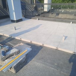 Stone MCh - Jasne płytki tarasowe ułożone na dachu budynku, widoczne narzędzia glazurnicze i poziomica. Częściowo widoczna osoba z telefonem.