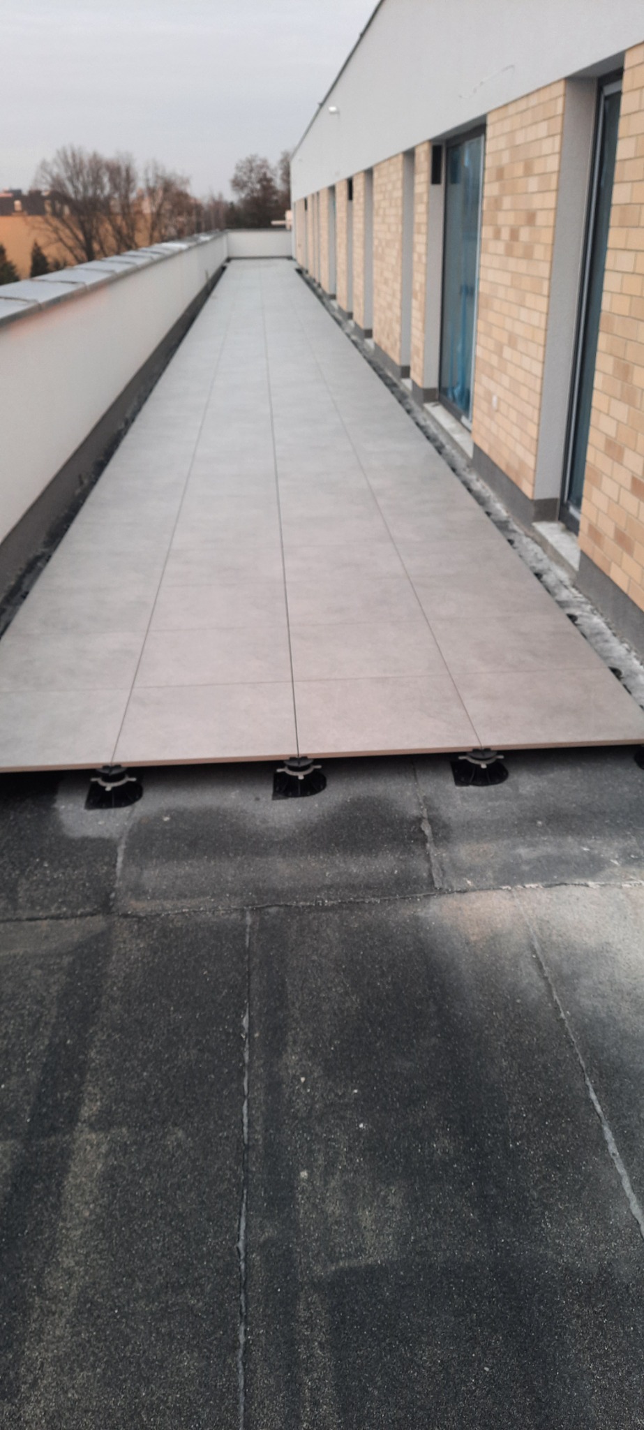 Montaż jasnoszarych płyt tarasowych na regulowanych wspornikach na dachu budynku z ceglaną elewacją i oknami. Widok z góry na ułożony fragment tarasu.