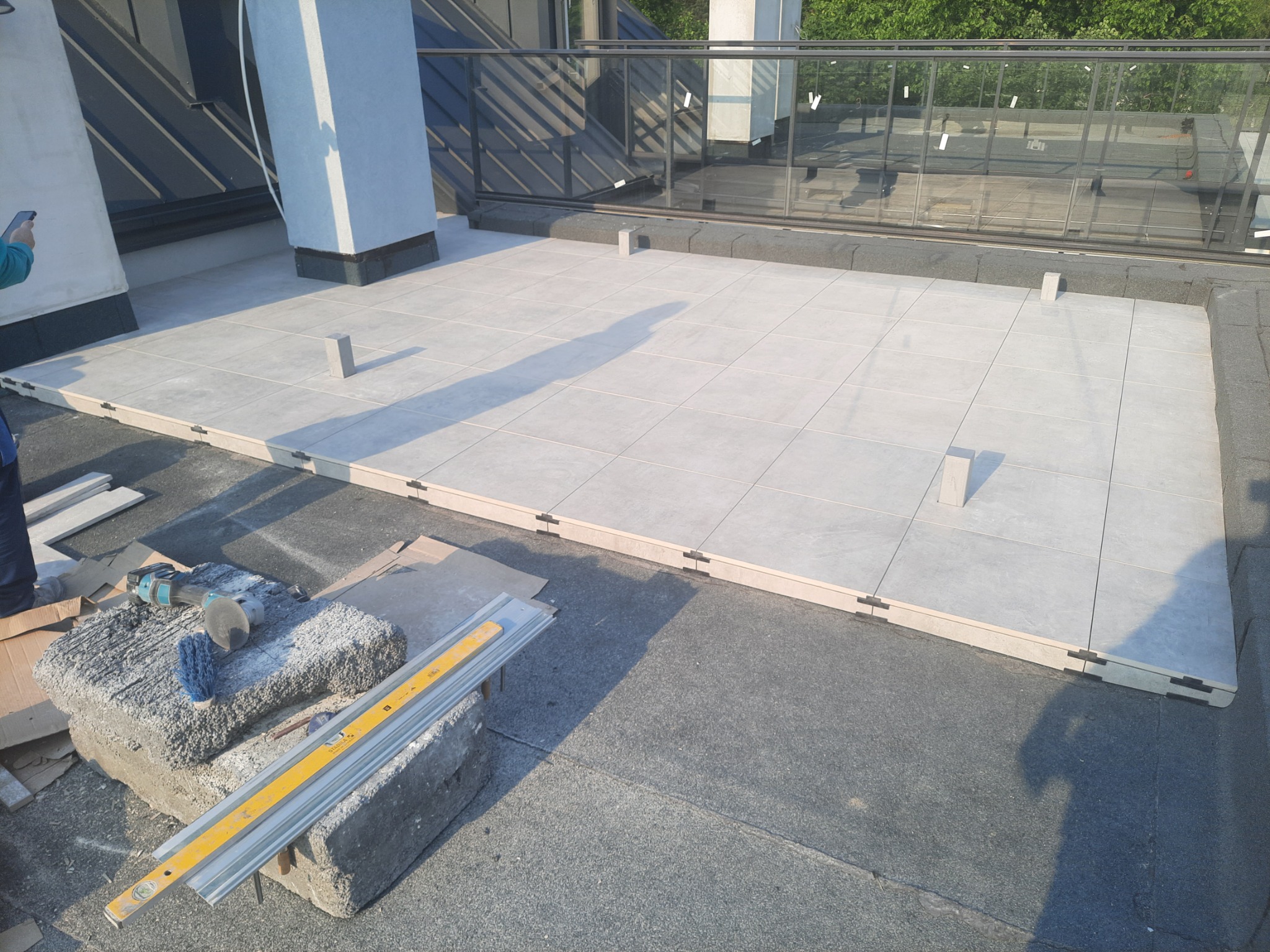 Jasne płytki tarasowe ułożone na dachu budynku, widoczne narzędzia glazurnicze i poziomica. Częściowo widoczna osoba z telefonem.