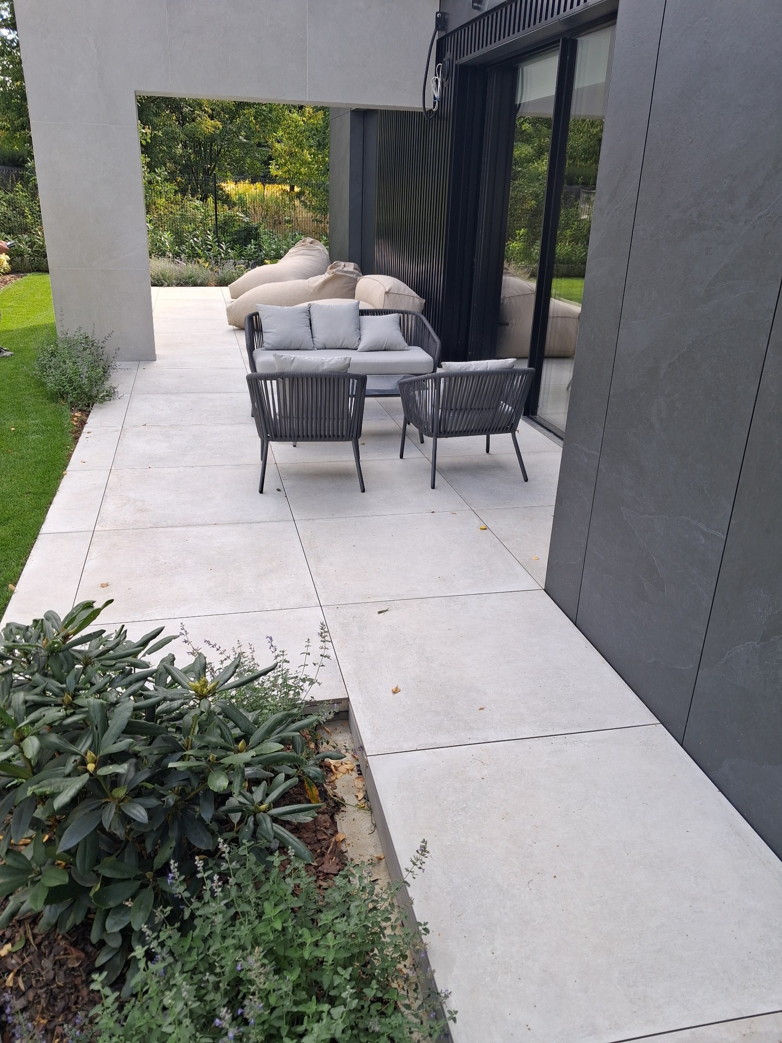 Nowoczesny taras z jasnych, kwadratowych płyt, szare meble ogrodowe, widok na zieleń. Minimalistyczny design, stonowana kolorystyka, eleganckie wykończenie.