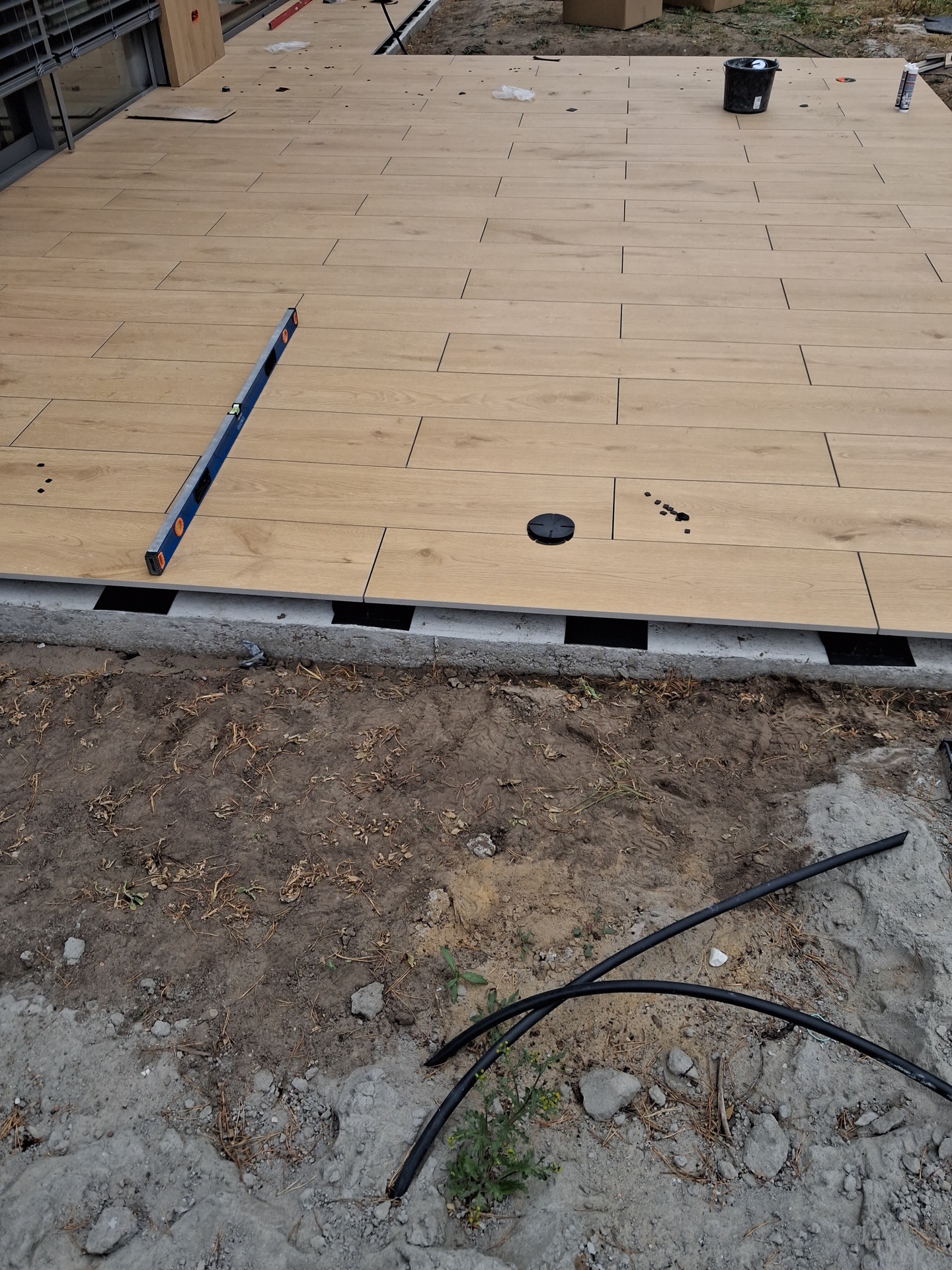 Układanie jasnych, drewnopodobnych płytek na tarasie. Widoczna poziomnica, wiadro i czarne rurki na ziemi. Częściowo ukończony taras.