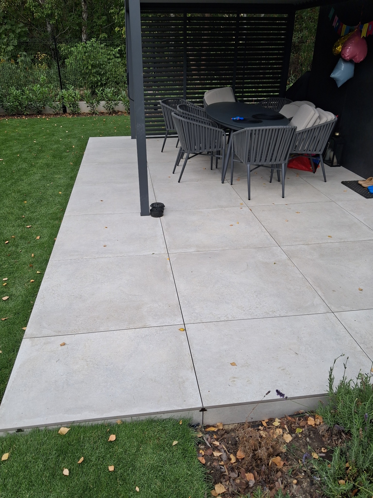Jasnoszary taras z płyt, z ciemnymi meblami ogrodowymi i zielonym trawnikiem. Widoczne detale wykończenia i roślinność.