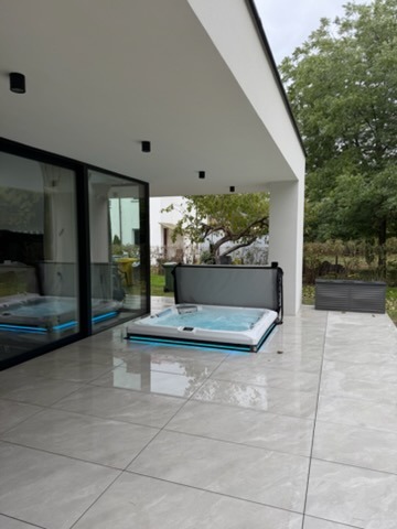 Nowoczesny taras z jasnoszarej kamiennej posadzki, na której stoi jacuzzi z podświetleniem LED i częściowo otwartą pokrywą. Widok na ogród zza szklanych drzwi.