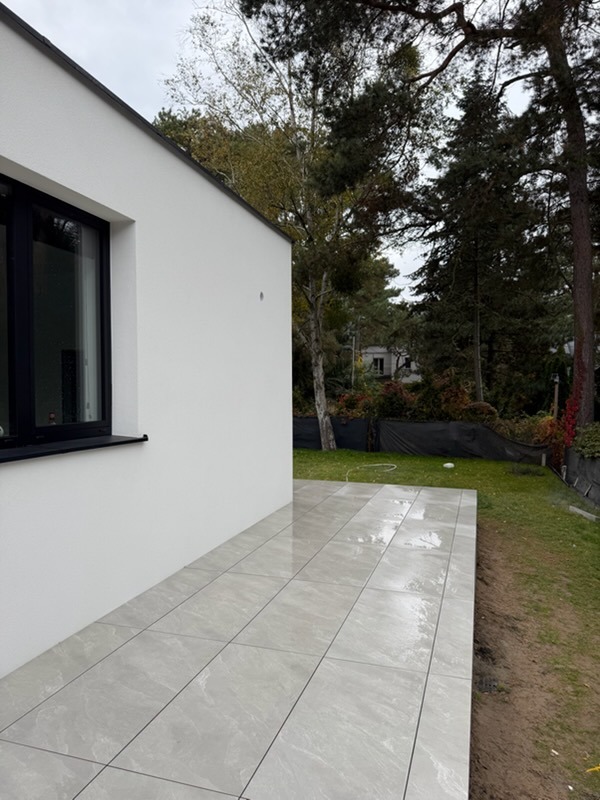 Jasnoszary taras z płytek ceramicznych przylegający do białej ściany budynku z czarnym oknem, widok na zieleń i drzewa w tle.