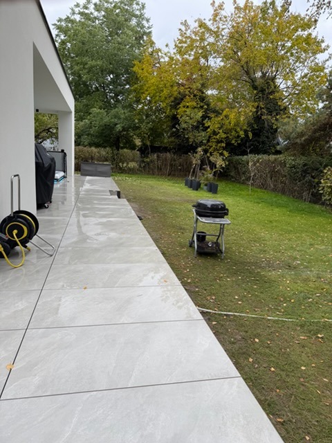 Nowoczesny taras z jasnych płyt w Warszawie, widok na ogród. Grill i narzędzia ogrodowe w tle. Minimalistyczny design z elementami zieleni.