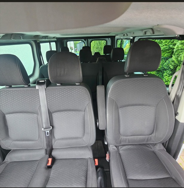 Wnętrze busa z rzędami foteli z zagłówkami i pasami bezpieczeństwa, widok z przodu pojazdu, okna z widokiem na zieleń.