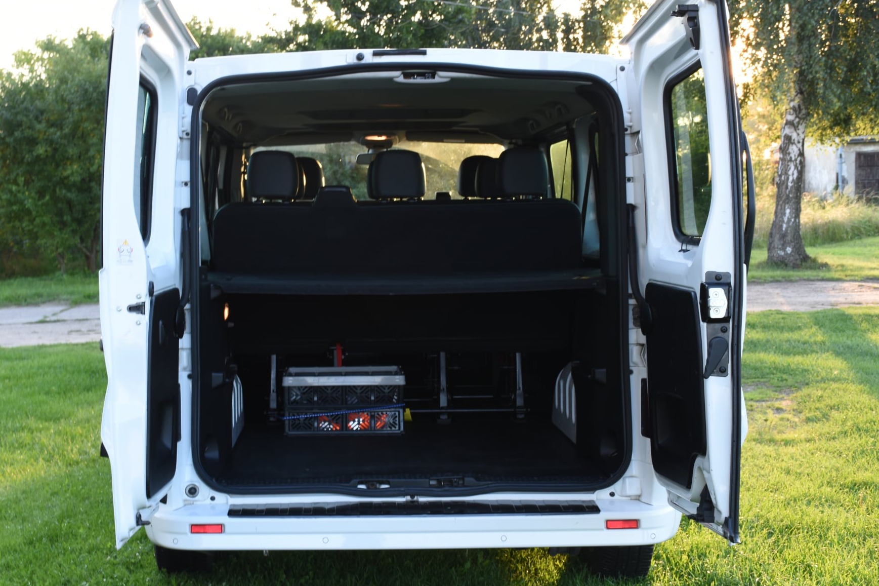 Wnętrze białego busa z otwartymi drzwiami z tyłu, widoczne siedzenia i bagażnik z kratką i wózkiem. Na zewnątrz trawa i drzewa.