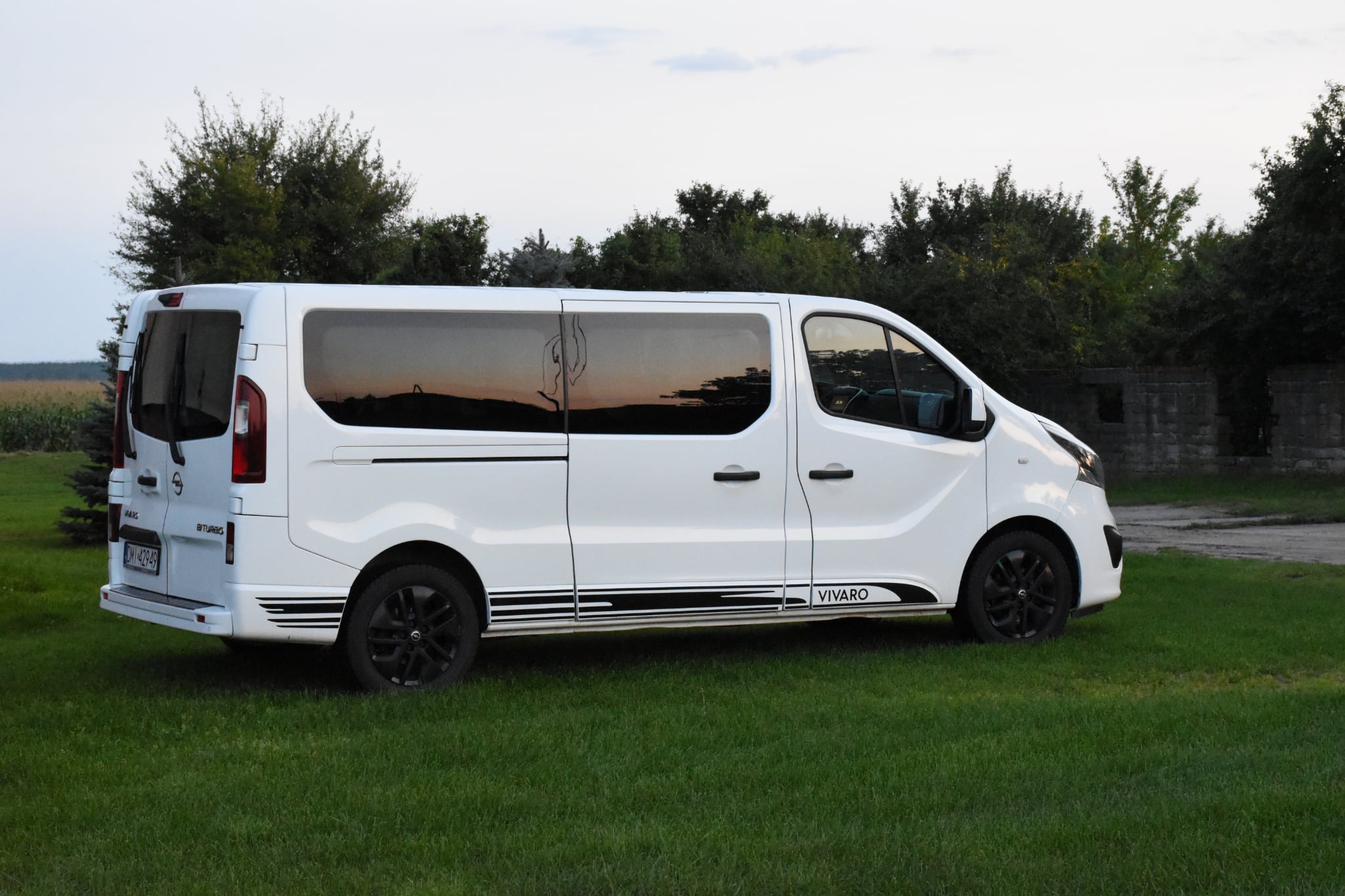 Biały bus Vivaro z czarnymi felgami i naklejkami na tle zielonej trawy i drzew. W oknach odbija się zachód słońca. Widok z boku, lekko od tyłu.