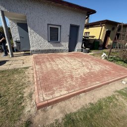 Andrzej Domin Uslugi Budowlane - Czerwona kostka brukowa ułożona na posesji przy budynku z oknem i szarymi drzwiami. Widoczny fragment trawnika i osoba w tle. Kostka zasypana piaskiem.