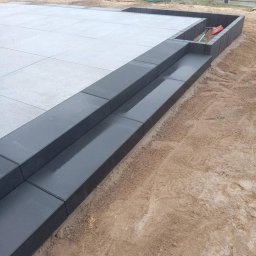 Andrzej Domin Uslugi Budowlane - Nowoczesny taras z jasnoszarej kostki, obrzeża z ciemnoszarej kostki, dwustopniowe schody. Widoczne narzędzia i kostka w opakowaniu. Tło: trawnik i ogrodzenie.