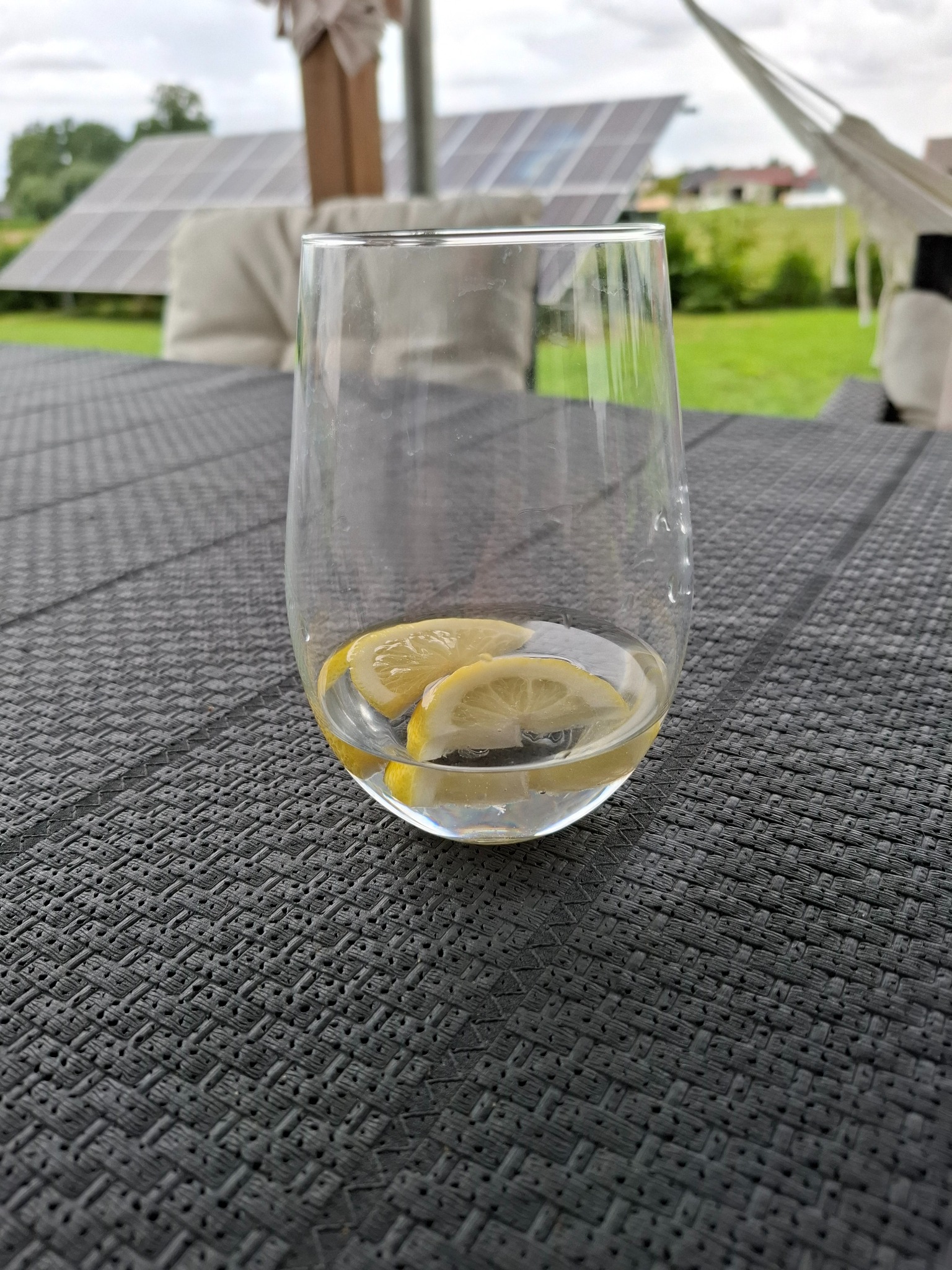 Szklanka z wodą i cytryną na tle panelu słonecznego, hamaka i zieleni. Letni relaks na tarasie.