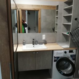 SYLWESTER PYSNO - Funkcjonalna łazienka w szarościach: umywalka nablatowa, szafki pod umywalką, pralka pod blatem i półki w zabudowie. W lustrze odbija się osoba robiąca zdjęcie.