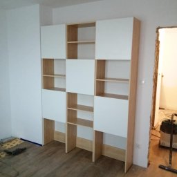 SYLWESTER PYSNO - Biało-drewniana szafa na wymiar z półkami, widziana w trakcie montażu. W tle widoczne narzędzia i przygotowane podłoże podłogowe. Oświetlenie dzienne.