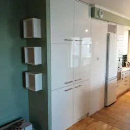 Biała, lakierowana zabudowa kuchenna z szafkami cargo i lodówką w tle, uzupełniona o trzy białe, wiszące półki na ścianie w odcieniach zieleni. Na podłodze panele.