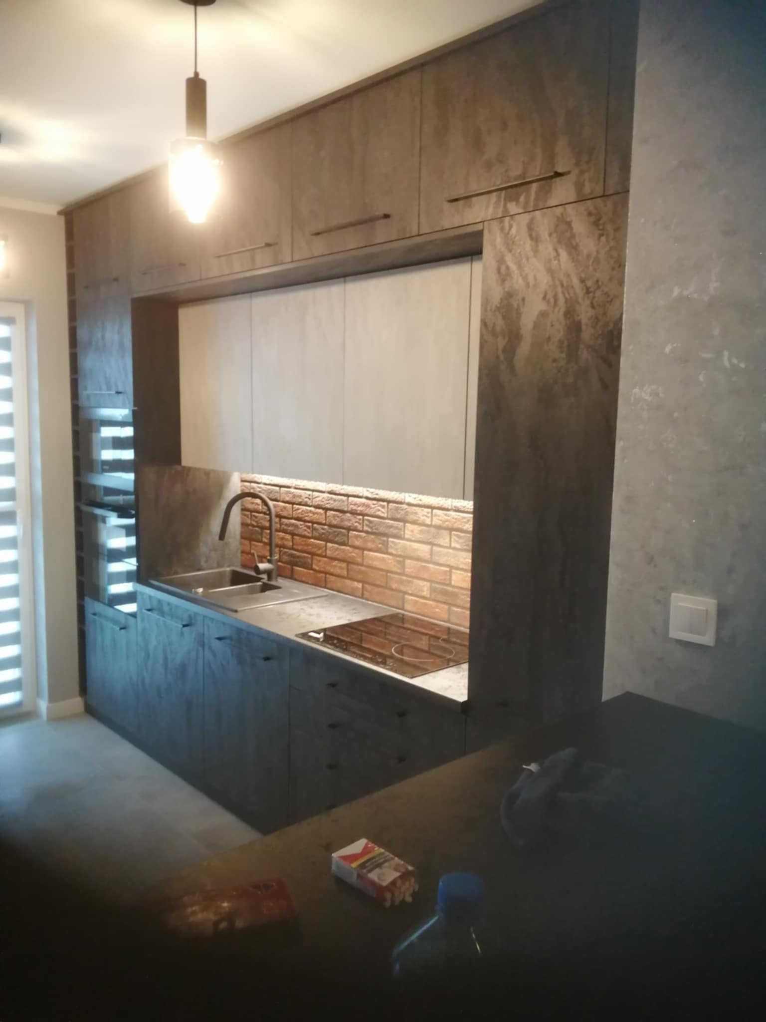 Nowoczesna kuchnia z szarymi szafkami, ceglaną ścianą nad blatem i minimalistycznym oświetleniem. Widoczne detale wykończenia i elementy wyposażenia.