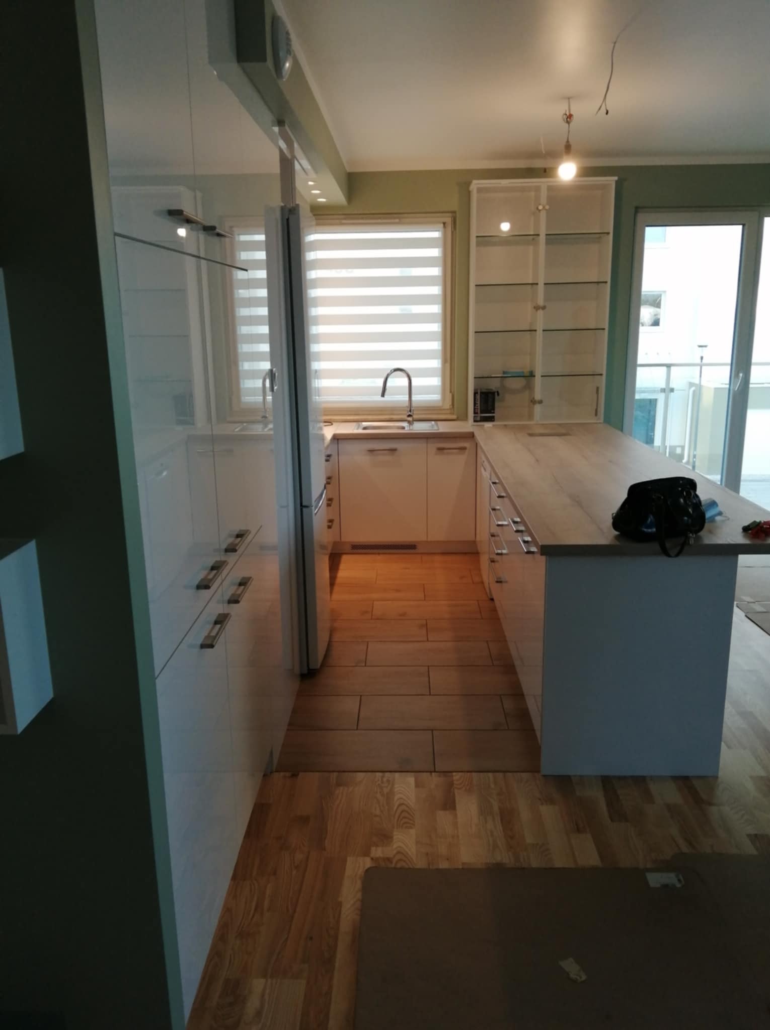 Jasna kuchnia z drewnianą podłogą i wyspą. Widoczne białe szafki z połyskiem i okno z żaluzjami. Na wyspie leży czarna torba. W tle balkon z przeszkleniem.