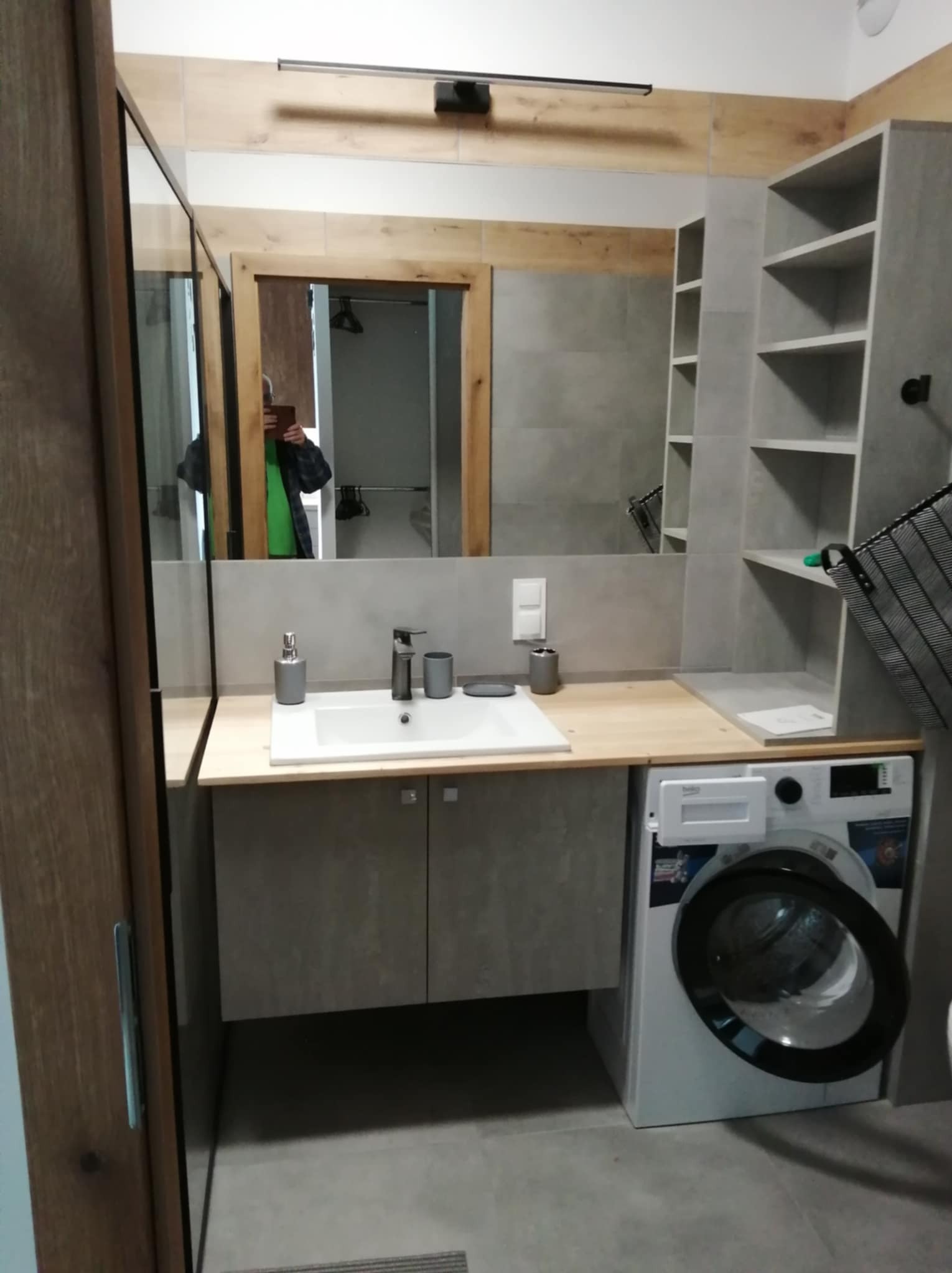 Funkcjonalna łazienka w szarościach: umywalka nablatowa, szafki pod umywalką, pralka pod blatem i półki w zabudowie. W lustrze odbija się osoba robiąca zdjęcie.