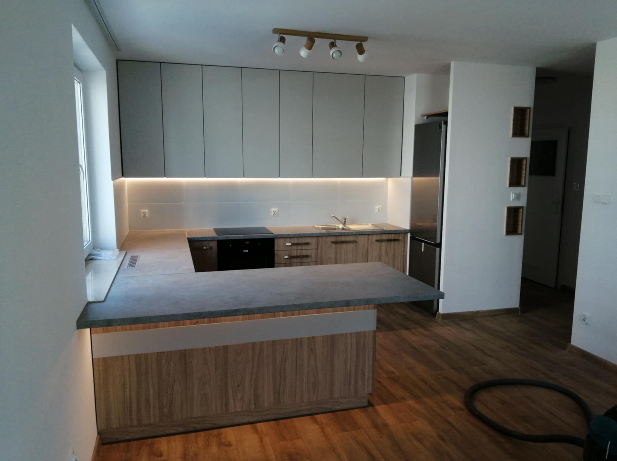 Nowoczesna kuchnia po remoncie: szare szafki wiszące z podświetleniem LED, drewniane fronty dolne, szary blat z wyspą. Widoczna podłoga z paneli i odkurzacz.